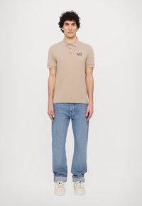 Beige polo skjorte med korte ermer og krage, med en liten EA7-logo. Kombinert med lyseblå jeans og hvite joggesko.