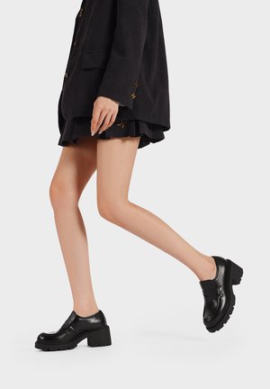 Scarpe platform in pelle nera con suola spessa, punta arrotondata, cinturino decorativo e dettagli di cucitura; texture liscia, design minimalista.