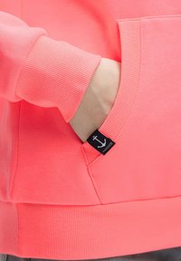 Coral sweatshirt with a ribbed hem and cuffs, featuring a front pocket and a small black tag with an anchor detail. Soft texture.