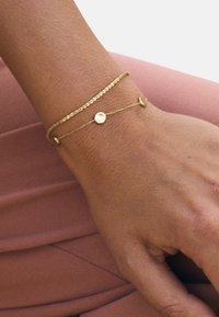 Złota bransoletka wielowarstwowa z teksturowanym łańcuszkiem i gładkim okrągłym elementem ozdobnym, spoczywająca na nadgarstku na różowym tle.