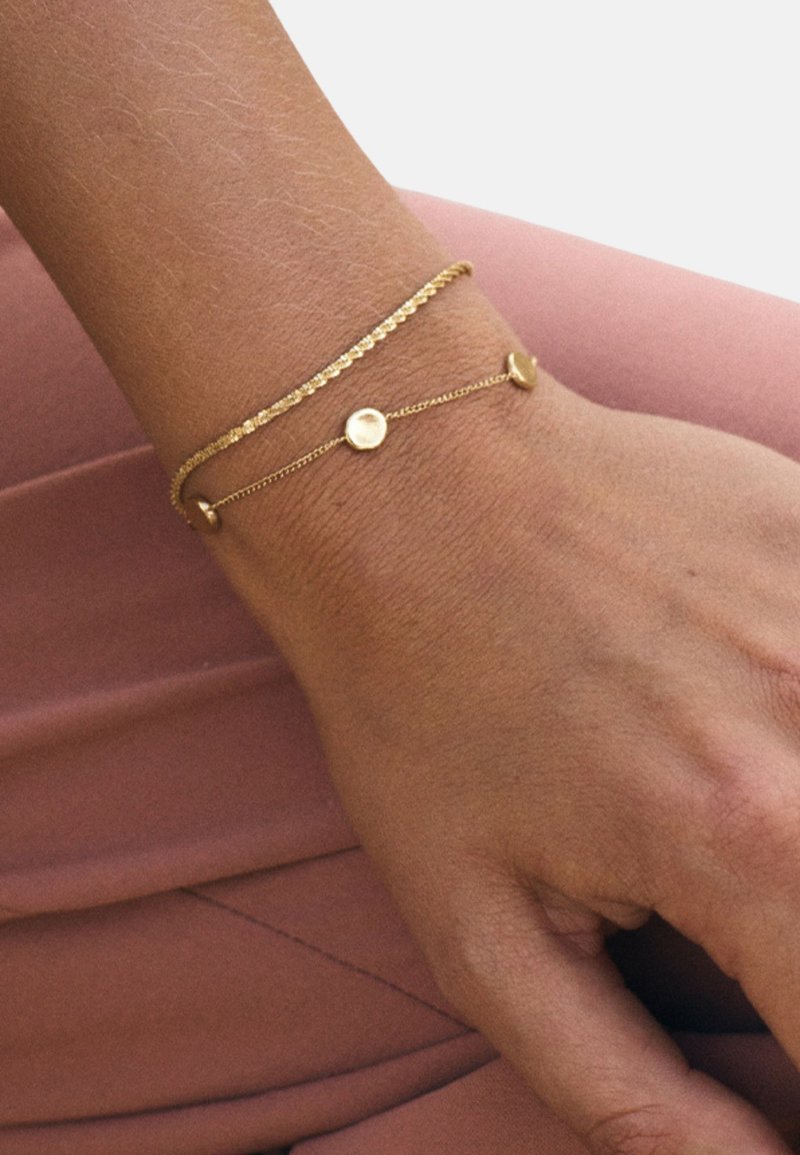 Złota bransoletka wielowarstwowa z teksturowanym łańcuszkiem i gładkim okrągłym elementem ozdobnym, spoczywająca na nadgarstku na różowym tle.