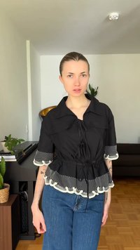 Γυναίκα με τα μαλλιά της χτενισμένα προς τα πίσω, φοράει μαύρο μπλουζάκι με λευκές κυματοειδείς άκρες και μπλε τζιν, στέκεται σε ένα σαλόνι με φυτά.