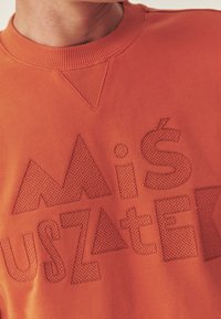 Orange sweatshirt med tekstureret broderi med geometriske former og bogstaver. Ribbet halsudskæring og en glat stoffinish.