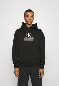 Felpa nera in cotone, dotata di una grande tasca frontale, cappuccio con cordino e logo ricamato Calvin Klein Jeans in colori a contrasto.