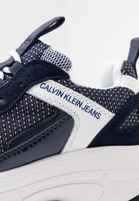 Marinblå sneakers i mesh och mocka, vita accenter, texturerad yta och "Calvin Klein Jeans"-logotyp på sidan.