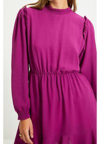 Robe fuchsia à manches longues, col froncé et poignets smockés. Taille élastique créant une silhouette fluide, réalisée dans un tissu léger.