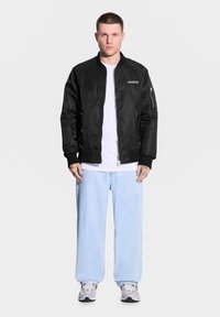 Czarna kurtka bomberka z zamkiem, ribbowane mankiety i naszywka na klatce piersiowej, noszona na białej koszuli i jasnoniebieskich luźnych spodniach z szarymi trampkami.