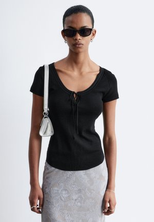 Femme portant un haut noir à manches courtes avec un nœud devant, une jupe fleurie argentée, des lunettes de soleil noires, un sac à épaule argenté et des bijoux minimalistes sur fond blanc.
