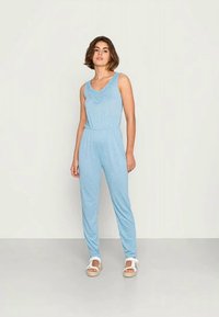 Macacão azul claro com decote texturizado, cintura elástica e corte descontraído, combinado com sandálias brancas sobre um fundo neutro.