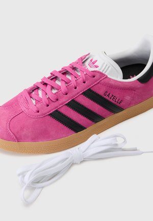 Różowy sneaker Adidas Gazelle z czarnymi paskami i gumową podeszwą, z różowymi sznurowadłami oraz dodatkowymi białymi sznurowadłami obok.