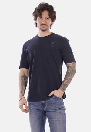 Uomo con capelli ricci che indossa una maglietta blu scuro della NYPD e jeans blu, mostrando tatuaggi dettagliati su entrambi gli avambracci, in posa con una mano in tasca.