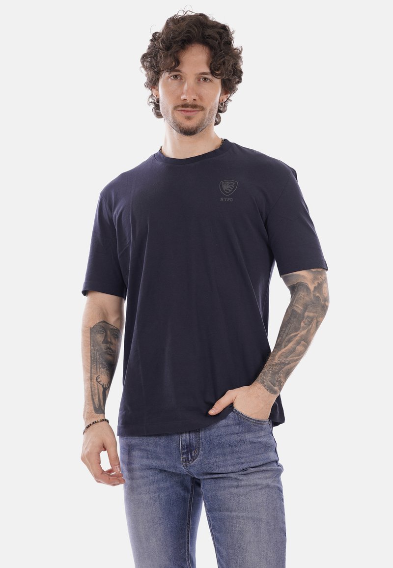 Uomo con capelli ricci che indossa una maglietta blu scuro della NYPD e jeans blu, mostrando tatuaggi dettagliati su entrambi gli avambracci, in posa con una mano in tasca.