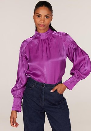 Paarse satijnen blouse met hoge hals, opgezette lange mouwen en gerimpelde details bij de schouders, gedragen met donkere denim jeans.