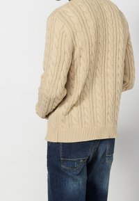 Pull beige en maille torsadée avec un motif texturé, doté d'un ourlet et de poignets côtelés. Associé à un jean bleu foncé légèrement usé.