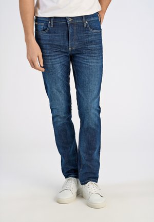 Dark blue denim jeans with a fitted cut, contrasting stitching, and five-pocket design. Paired with white sneakers.