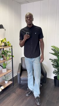 Black button-up shirt, light blue jeans, and dark shoes. Standing in a well-lit room with plants and a black chair.