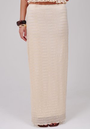 Femme portant une jupe longue texturée crème avec un haut à volants, des bracelets marron et beige au poignet gauche, et des sandales marron à bouts ouverts.