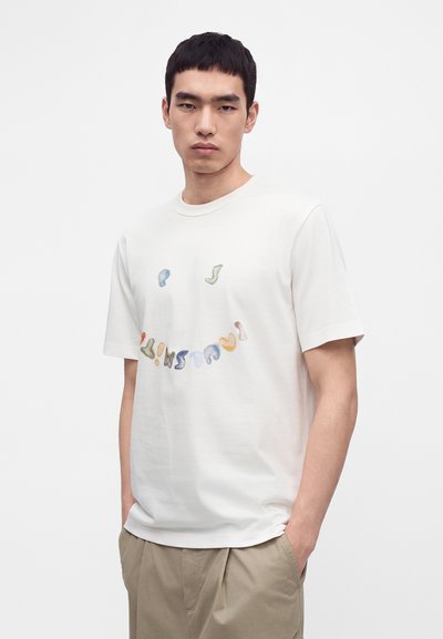 Νέος άνδρας που φοράει λευκό T-shirt με πολύχρωμο σχέδιο χαμογελαστής φατσούλας, στέκεται με τα χέρια στις τσέπες μπεζ παντελονιού μπροστά σε απλό φόντο.