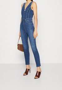 Mono de mezclilla con escote en V y silueta ajustada. Presenta detalles de costura y cintura ceñida con cinturón. Combinado con mules de tacón marrones.