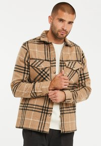 Tan geruite shirtjack met zwart-wit ruitpatroon, twee borstzakken en knoopsluiting. Gemaakt van zacht, warm materiaal.