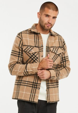 Tan geruite shirtjack met zwart-wit ruitpatroon, twee borstzakken en knoopsluiting. Gemaakt van zacht, warm materiaal.