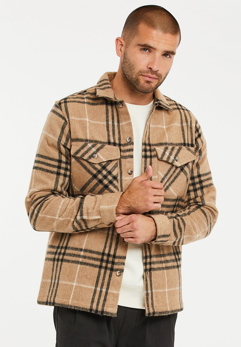 Tan geruite shirtjack met zwart-wit ruitpatroon, twee borstzakken en knoopsluiting. Gemaakt van zacht, warm materiaal.