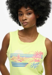 Fel geel mouwloos topje met een grafisch ontwerp van een blauwe en roze surfer, palmbomen en zon tegen horizontale strepen.