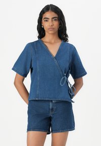 Top de denim com mangas curtas, fecho com laço na cintura, e uma cor azul sólida com um acabamento limpo e detalhes de costura.