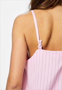 Η άνω πλάτη γυναίκας που φοράει ροζ αμάνικο τοπ με ρίγες και δεμένο ιμάντα πάνω σε λευκό απλό φόντο.
