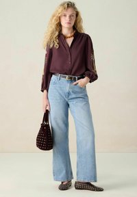 Bordeauxrode blouse met geborduurde mouwen, lichtblauwe broek met wijde pijpen, bruine platte schoenen met studs, en een bordeauxrode emmertas met oogjes.