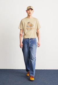 Beige graphic T-shirt featuring Timberland logo, blue denim jeans, and colorful sneakers, complemented by a matching cap.