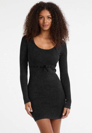 Woman with curly hair wearing a fitted black ribbed long-sleeve dress with scoop neckline and small bow detail at the waist.