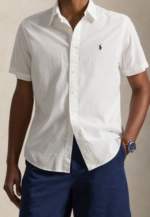 Homme portant une chemise blanche à manches courtes avec un petit logo brodé bleu, un pantalon bleu marine et une montre-bracelet marron.