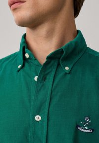 Grünes Cordhemd mit Button-Down-Kragen, weißen Knöpfen und einem gestickten Logo auf der Brust. Strukturierter Stoff mit vertikalen Streifen.