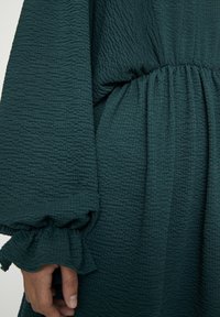 Robe verte texturée avec une taille élastique, des manches bouffantes et des poignets à volants. Le tissu présente un motif froissé pour plus de détails.