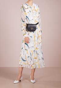 Robe à manches longues avec un motif floral en blanc, jaune et teal ; design à volants avec un sac ceinture noir, associée à des escarpins blancs à bout pointu.