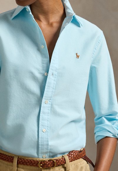 Person trägt hellblaues Hemd mit Knöpfen und hochgekrempelten Ärmeln, beige Hose und geflochtenen braunen Gürtel mit gesticktem Logo auf der Brust.