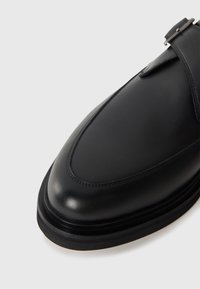 Black leather dress shoe with a rounded toe, smooth texture, single strap design with a metallic buckle, and a layered rubber sole.