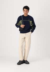 Sweat-shirt marine avec des accents verts et gris, associé à un pantalon beige clair. Le modèle porte des chaussures noires et un sac noir. Design minimaliste.
