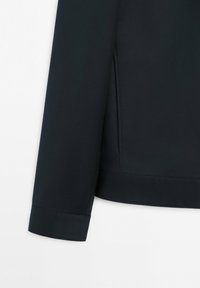 Manica e cucitura laterale di una giacca blu navy scuro con un piccolo dettaglio a spacco, appoggiata su uno sfondo bianco.