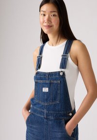 Denimoverall i mörkblått, med justerbara axelremmar, framficka och sidoknappar. Bärs över en vit linne.