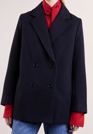 Personne portant un manteau bleu marine croisé sur une blouse rouge avec de larges poignets et un jean bleu clair, posant devant un fond uni.