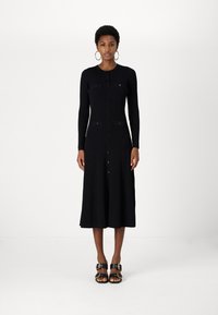 Fekete bordázott kötött ruha hosszú ujjakkal és gombos elülső résszel. Két felvarrt zsebbel rendelkezik, midi hosszúságú. Fekete szandálokkal viselik.