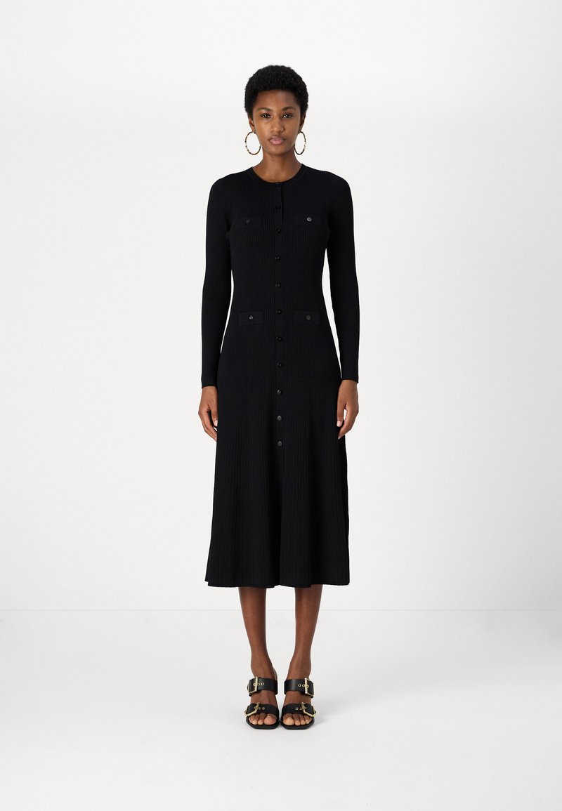 Fekete bordázott kötött ruha hosszú ujjakkal és gombos elülső résszel. Két felvarrt zsebbel rendelkezik, midi hosszúságú. Fekete szandálokkal viselik.