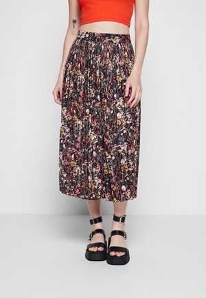 Fließender Midirock in Schwarz mit floralen Mustern, die rosa, gelbe und weiße Blumen zeigen; elastischer Taillenbund; kombiniert mit klobigen schwarzen Sandalen.
