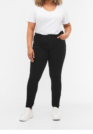 Femme portant un jean skinny noir, un t-shirt blanc à manches courtes et des baskets blanches, debout avec une main dans la poche sur un fond blanc.