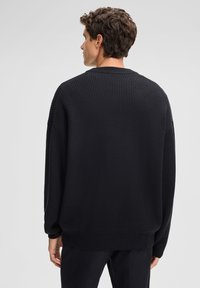 Uomo con capelli ricci corti che indossa un maglione nero a coste largo, in piedi girato verso un semplice sfondo grigio chiaro.