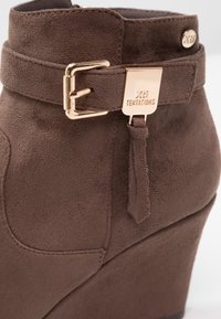 Stivaletto in suede marrone con tacco a zeppa, dotato di una fibbia dorata e una placca con logo, e un dettaglio decorativo con cinturino e nappina.