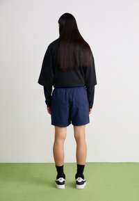 En person med långt mörkt hår står vänd mot en vit vägg, klädd i en svart topp, marinblå shorts, svarta strumpor och vita Nike-sneakers.