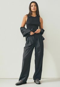 Zwarte geribbelde tanktop gecombineerd met losse zwarte broeken, Accentueert door een gedrapeerde zwarte jas. Draagt met gestructureerde zwarte loafers.